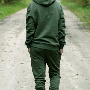 Pu&ograve; includere: Una persona indossa una felpa con cappuccio verde e pantaloni della tuta abbinati mentre cammina su un sentiero sterrato. Indossa scarpe da ginnastica bianche.