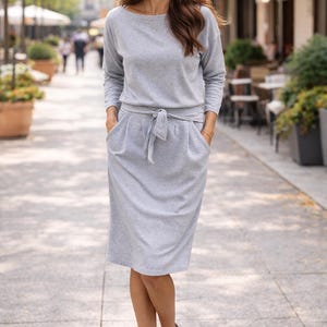 May include: A light gray dress with long sleeves, a tie waist, and pockets. The dress is paired with black heels. The woman is walking on a paved walkway with trees and buildings in the background.
