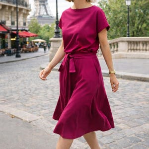 May include: A woman wearing a magenta-colored dress with a tie waist and short sleeves. The dress is paired with white slip-on shoes. The Eiffel Tower is visible in the background, suggesting a European setting.