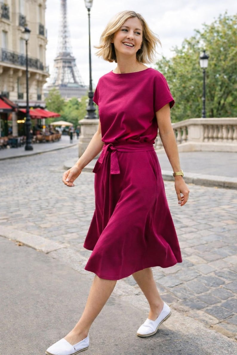 May include: A woman wearing a magenta-colored dress with a tie waist and short sleeves. The dress is paired with white slip-on shoes. The Eiffel Tower is visible in the background, suggesting a European setting.