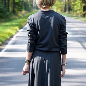 May include: A woman wearing a long, gray,  sleeveless dress with a small cross necklace. She is walking down a paved road with trees on either side.