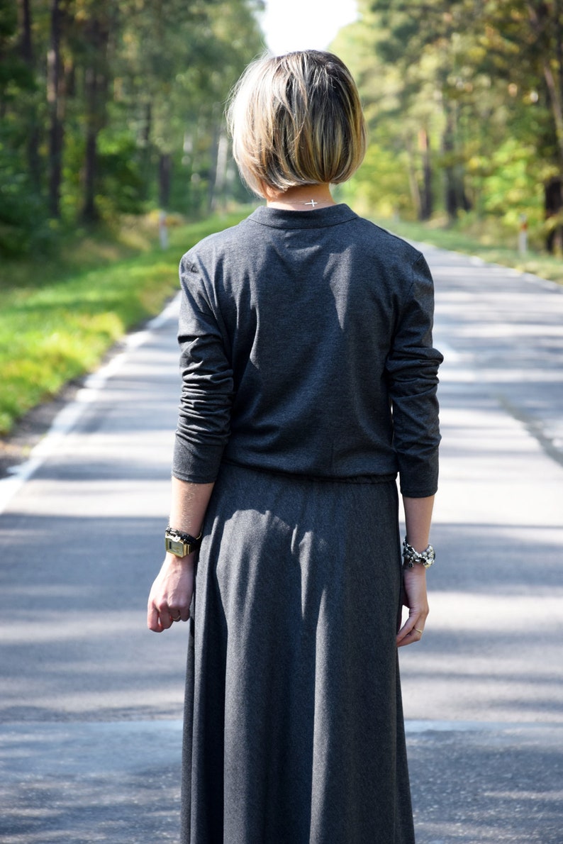 May include: A woman wearing a long, gray,  sleeveless dress with a small cross necklace. She is walking down a paved road with trees on either side.