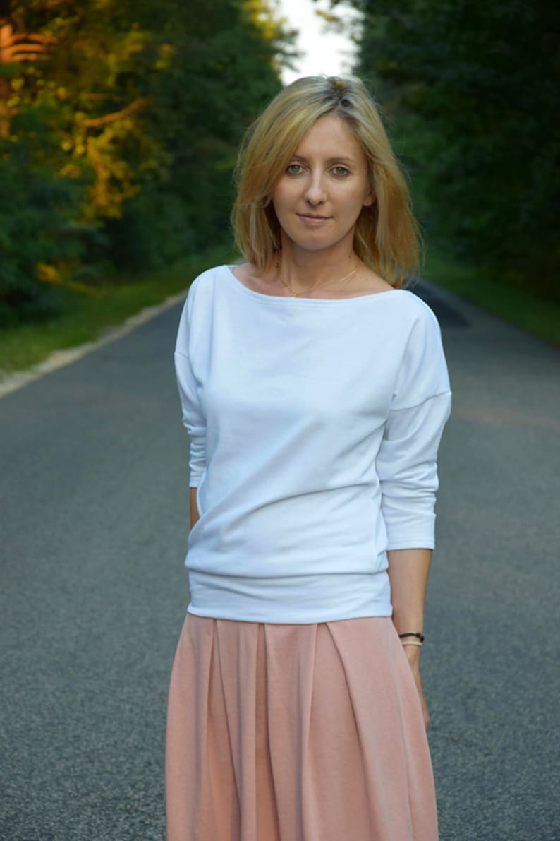 Puede incluir: Una mujer con una camisa blanca de manga larga y una falda plisada de color melocot&oacute;n. Est&aacute; de pie en un camino pavimentado con &aacute;rboles a ambos lados.