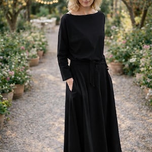 May include: A black midi dress with long sleeves and a boat neckline. The dress features a tie waist and pockets. The person is wearing black heels. The background shows a garden path with potted plants and trees.