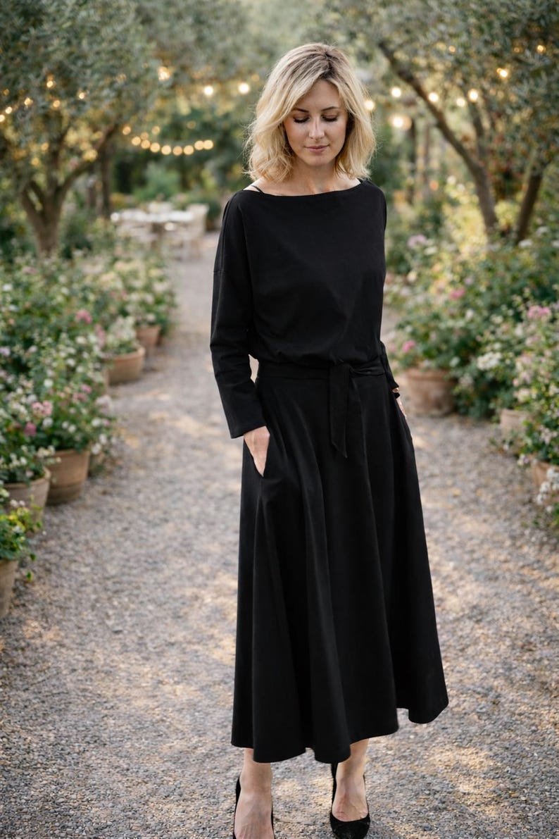 May include: A black midi dress with long sleeves and a boat neckline. The dress features a tie waist and pockets. The person is wearing black heels. The background shows a garden path with potted plants and trees.