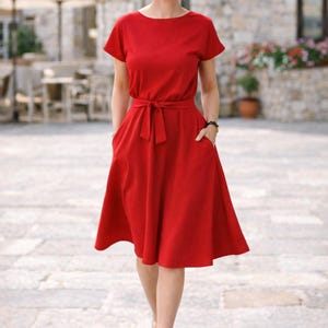 May include: A red dress with short sleeves, a tie waist, and pockets. The dress is knee-length and is worn with white heels. The background features a stone building and outdoor seating.