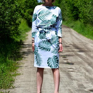 May include: A white knee-length dress featuring a green tropical palm leaf print, three-quarter length sleeves, a boat neckline, and a matching fabric tie belt at the waist. The garment is paired with light gray pointed-toe high heel shoes.