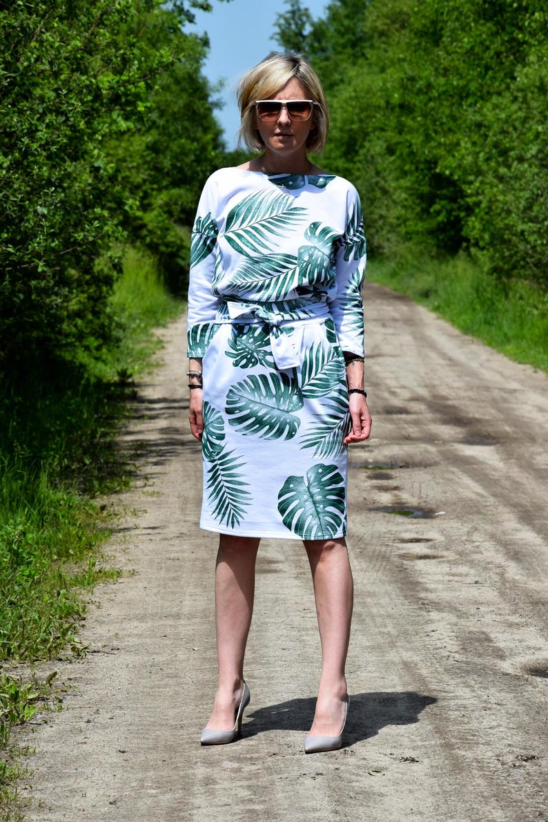 May include: A white knee-length dress featuring a green tropical palm leaf print, three-quarter length sleeves, a boat neckline, and a matching fabric tie belt at the waist. The garment is paired with light gray pointed-toe high heel shoes.