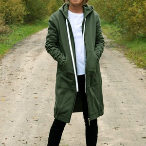 May include: A woman wearing a long, olive green hooded sweatshirt with a white zipper and white drawstrings. She is wearing black pants and white sneakers with pink accents.