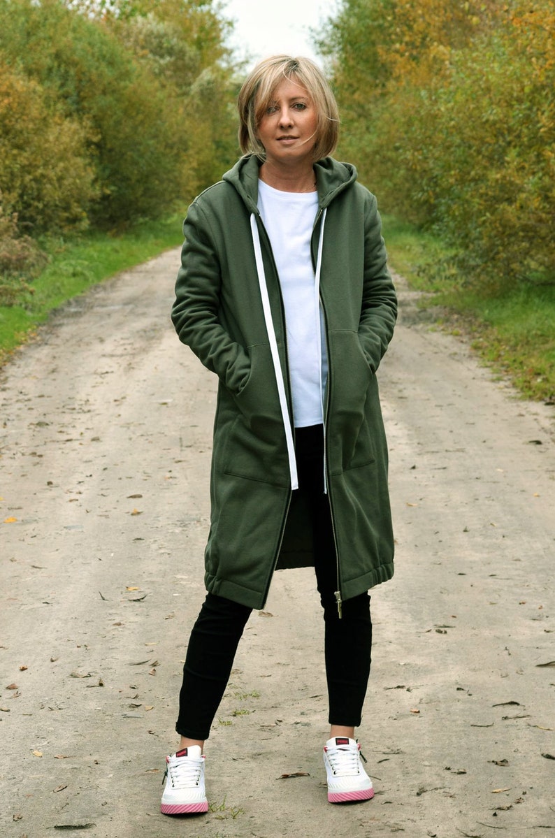 May include: A woman wearing a long, olive green hooded sweatshirt with a white zipper and white drawstrings. She is wearing black pants and white sneakers with pink accents.