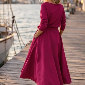 May include: A magenta-coloured dress with long sleeves and a flowing skirt, cinched at the waist with a matching belt. The dress has a scoop neckline and pockets. The person is wearing white trainers. The background shows a wooden pier and water.