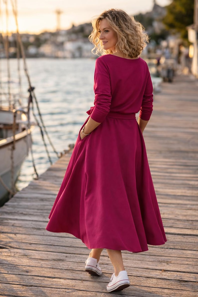 May include: A magenta-coloured dress with long sleeves and a flowing skirt, cinched at the waist with a matching belt. The dress has a scoop neckline and pockets. The person is wearing white trainers. The background shows a wooden pier and water.