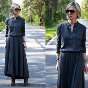 May include: A woman wearing a long, gray, collared dress with long sleeves and a high neckline. She is wearing brown boots and sunglasses. The dress has a pocket on the left side. She is standing on a paved road with trees on either side.