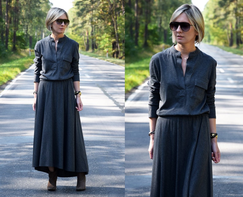 May include: A woman wearing a long, gray, collared dress with long sleeves and a high neckline. She is wearing brown boots and sunglasses. The dress has a pocket on the left side. She is standing on a paved road with trees on either side.