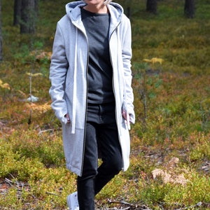 May include: A woman wearing a long, gray hooded sweatshirt with a zipper and black jeans. She is walking in a forest and wearing white sneakers.