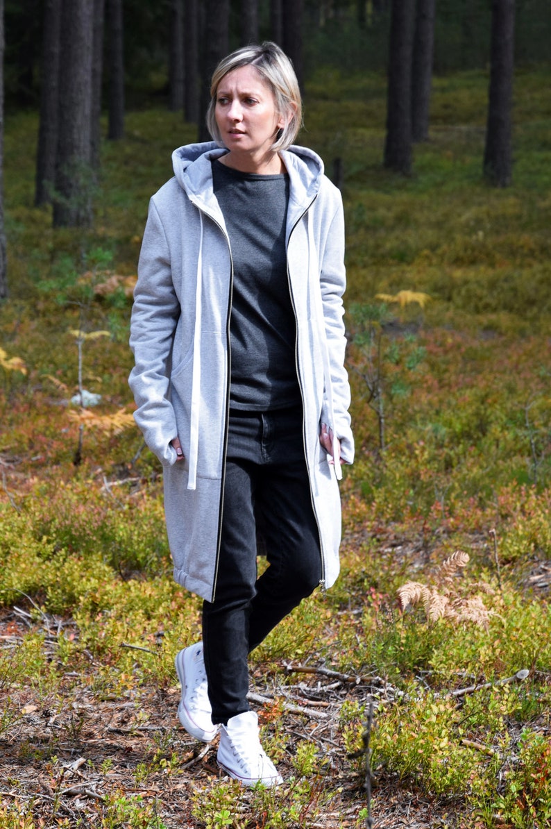May include: A woman wearing a long, gray hooded sweatshirt with a zipper and black jeans. She is walking in a forest and wearing white sneakers.