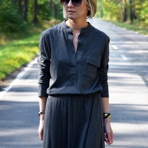 May include: A woman wearing a long, gray, collared dress with a pocket on the left side. She is wearing sunglasses and a silver necklace.