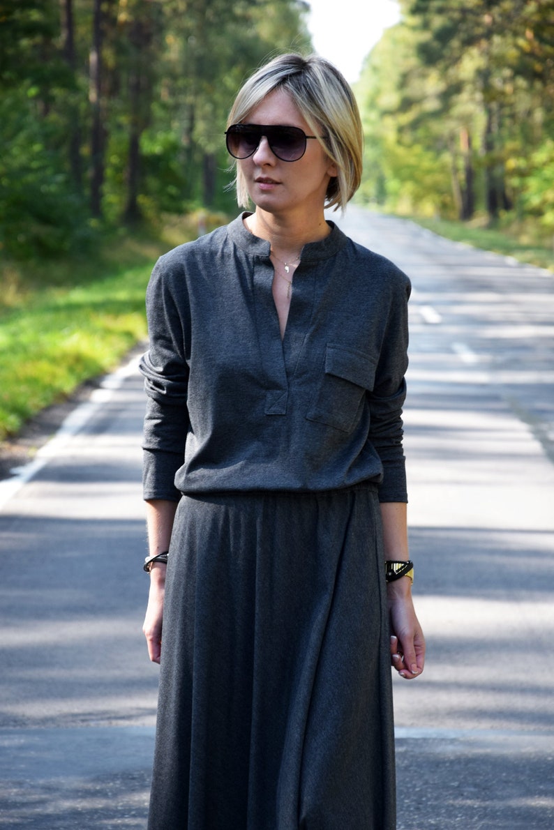 May include: A woman wearing a long, gray, collared dress with a pocket on the left side. She is wearing sunglasses and a silver necklace.