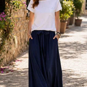 Könnte beinhalten: Eine Frau trägt ein weißes T-Shirt und eine marineblaue Hose mit weitem Bein und Bindeband. Das Outfit ist lässig und bequem, passend für warmes Wetter. Sie geht auf einer gepflasterten Straße.