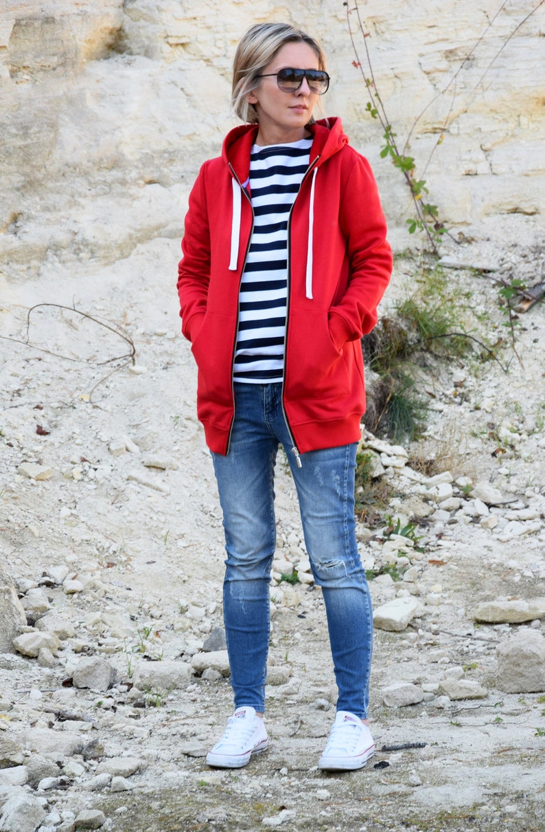 Pode incluir: Uma mulher vestindo um moletom vermelho com capuz e z&iacute;per, uma camisa listrada, jeans azul e t&ecirc;nis branco. Ela est&aacute; em uma &aacute;rea rochosa.