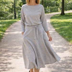 May include: A pale grey dress with long sleeves and a tie waist. The dress features a boat neck and a flowing skirt. The person is wearing black heels and walking on a path in a park.