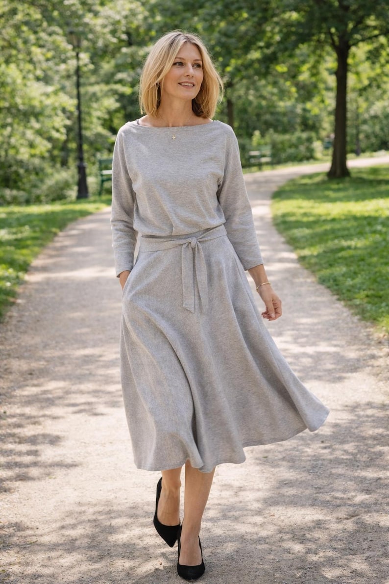 May include: A pale grey dress with long sleeves and a tie waist. The dress features a boat neck and a flowing skirt. The person is wearing black heels and walking on a path in a park.