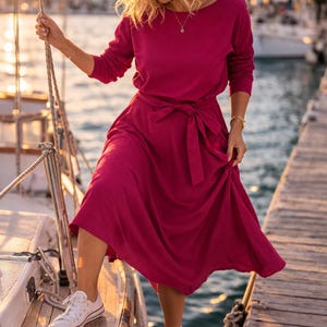 May include: A magenta dress with a tie waist and long sleeves, worn with white trainers. The dress features a boat neckline. The scene is set on a wooden dock next to a sailboat, with a coastal backdrop.