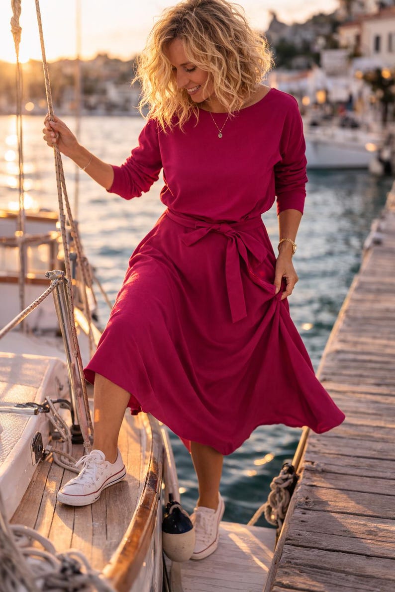 May include: A magenta dress with a tie waist and long sleeves, worn with white trainers. The dress features a boat neckline. The scene is set on a wooden dock next to a sailboat, with a coastal backdrop.