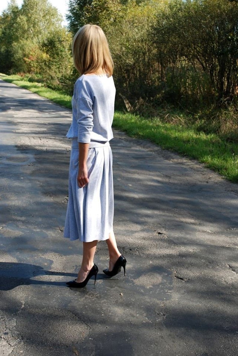 Puede incluir: Una mujer con un vestido de su&eacute;ter gris claro y tacones altos negros camina por un camino pavimentado.