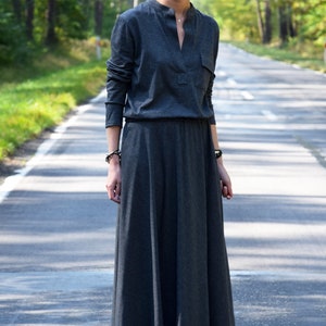 May include: A woman wearing a long, dark gray maxi dress with a v-neck and long sleeves. She is wearing sunglasses and brown ankle boots.