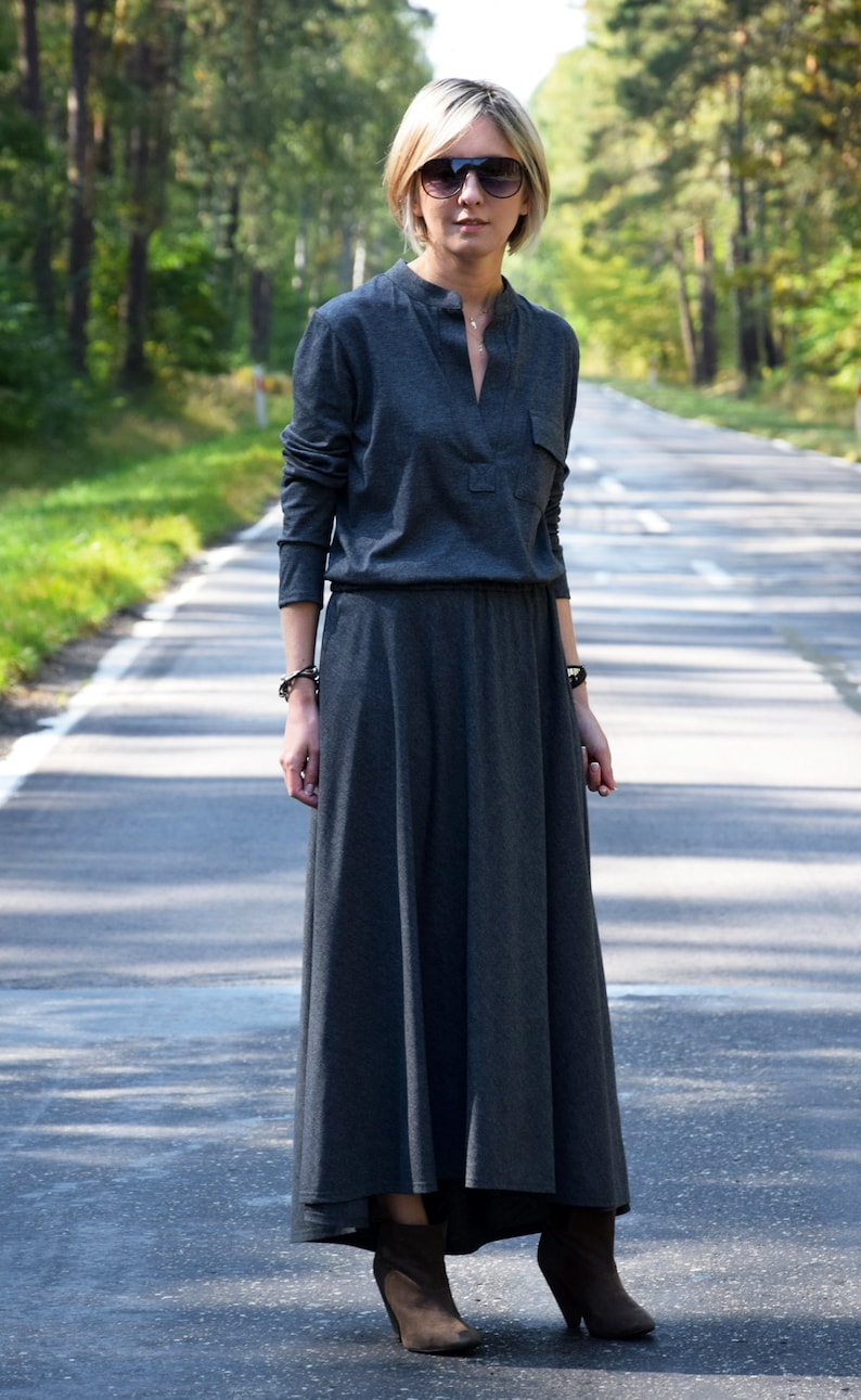 May include: A woman wearing a long, dark gray maxi dress with a v-neck and long sleeves. She is wearing sunglasses and brown ankle boots.