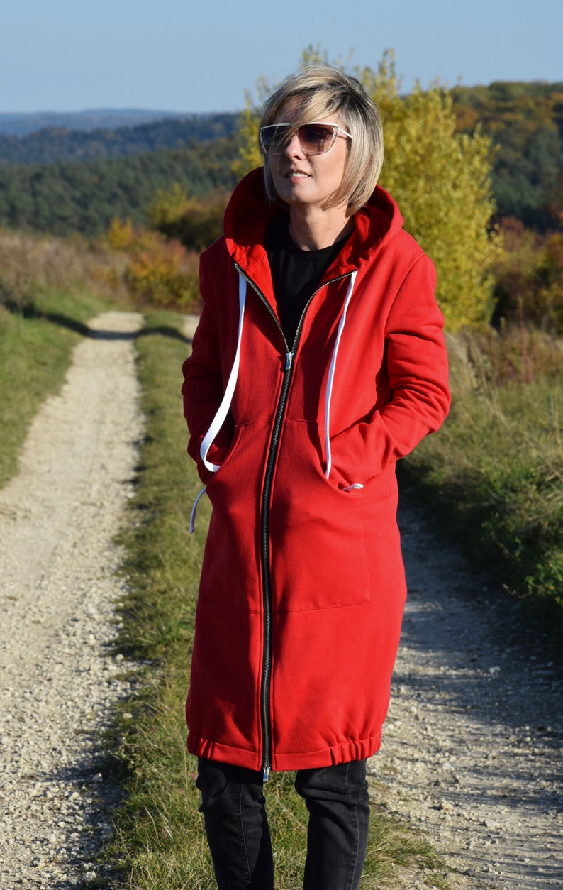 May include: A long red hooded sweatshirt with a black zipper and white drawstrings. The sweatshirt has a relaxed fit and a slightly gathered hem.