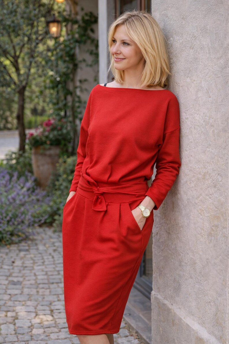 May include: A red dress with long sleeves, a boat neckline, and a tie-waist detail. The dress has pockets and is knee-length. A gold wristwatch is visible on the wrist.