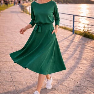 May include: A woman in a long-sleeved, emerald green dress with a tie waist and pockets, and white trainers. The dress features a boat neck and a flowing skirt. The background shows a waterfront promenade.