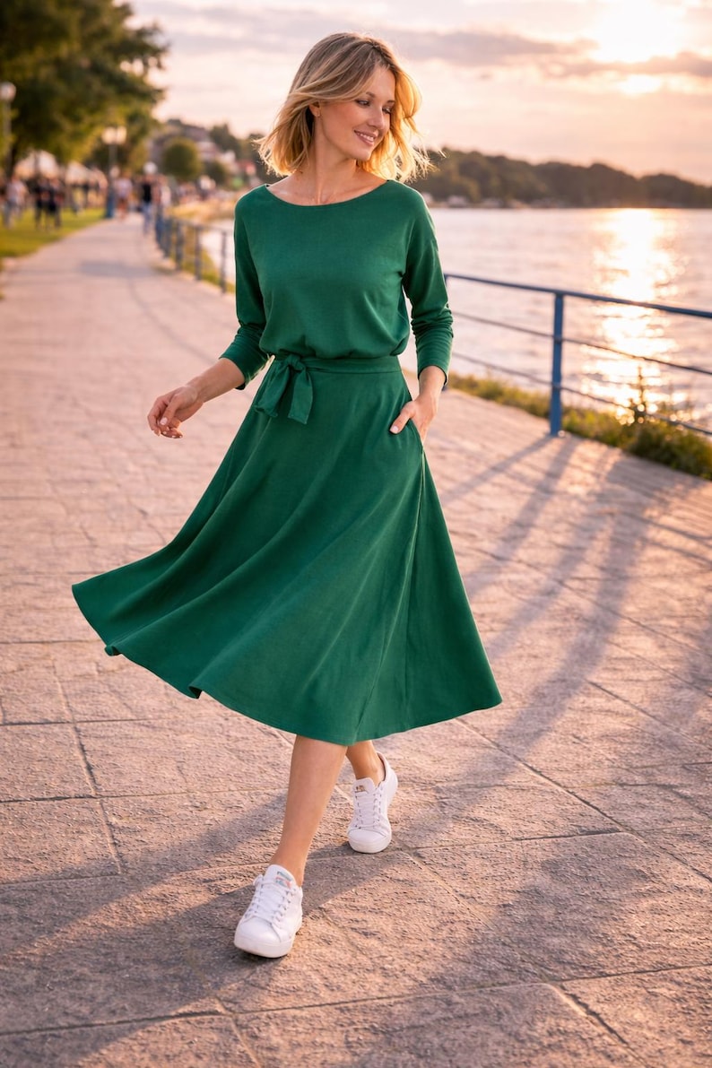 May include: A woman in a long-sleeved, emerald green dress with a tie waist and pockets, and white trainers. The dress features a boat neck and a flowing skirt. The background shows a waterfront promenade.