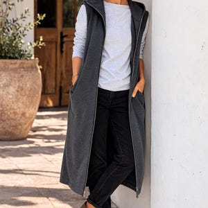 Puede incluir: Una mujer con un chaleco largo con capucha gris oscuro con cremallera completa, sobre una camiseta de manga larga gris claro y vaqueros negros. Lleva botines marrones y gafas de sol. El atuendo es informal y elegante.