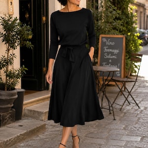 May include: A black dress with a boat neckline and a tie waist. The dress has long sleeves and a flared skirt. The person is wearing black sandals. The background includes a restaurant with a chalkboard sign.