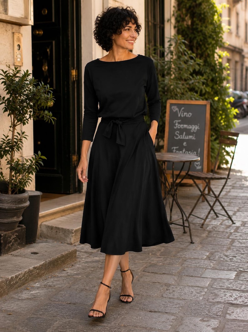 May include: A black dress with a boat neckline and a tie waist. The dress has long sleeves and a flared skirt. The person is wearing black sandals. The background includes a restaurant with a chalkboard sign.