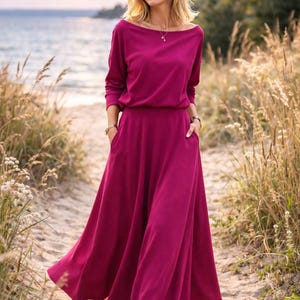 May include: A long, magenta dress with a boat neckline and long sleeves. The dress has a fitted waist and a flowing skirt with pockets. The person is wearing white sneakers. The background includes a beach and tall grass.