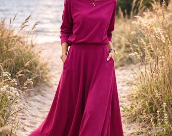 May include: A long, magenta dress with a boat neckline and long sleeves. The dress has a fitted waist and a flowing skirt with pockets. The person is wearing white sneakers. The background includes a beach and tall grass.
