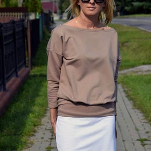 Puede incluir: Una mujer con una blusa marr&oacute;n de manga larga y holgada con una falda blanca. Lleva gafas de sol negras.