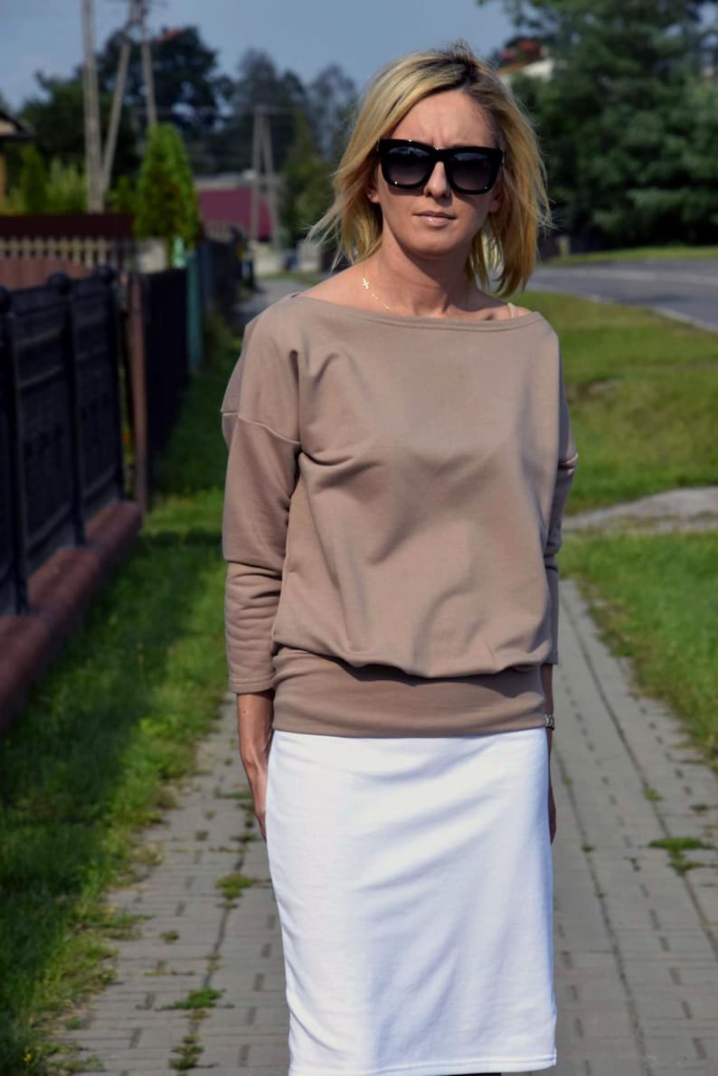 Puede incluir: Una mujer con una blusa marr&oacute;n de manga larga y holgada con una falda blanca. Lleva gafas de sol negras.