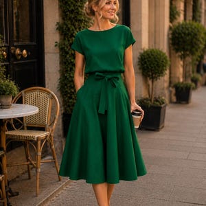 May include: A woman wearing a green dress with a matching belt and short sleeves, holding a coffee cup. The dress has a flared skirt and pockets. She is wearing beige heels and is walking on a sidewalk.