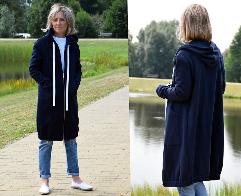 May include: A navy blue hooded sweatshirt with a zipper closure and white drawstrings. The sweatshirt has a long, relaxed fit and is shown being worn by a person in a park setting.