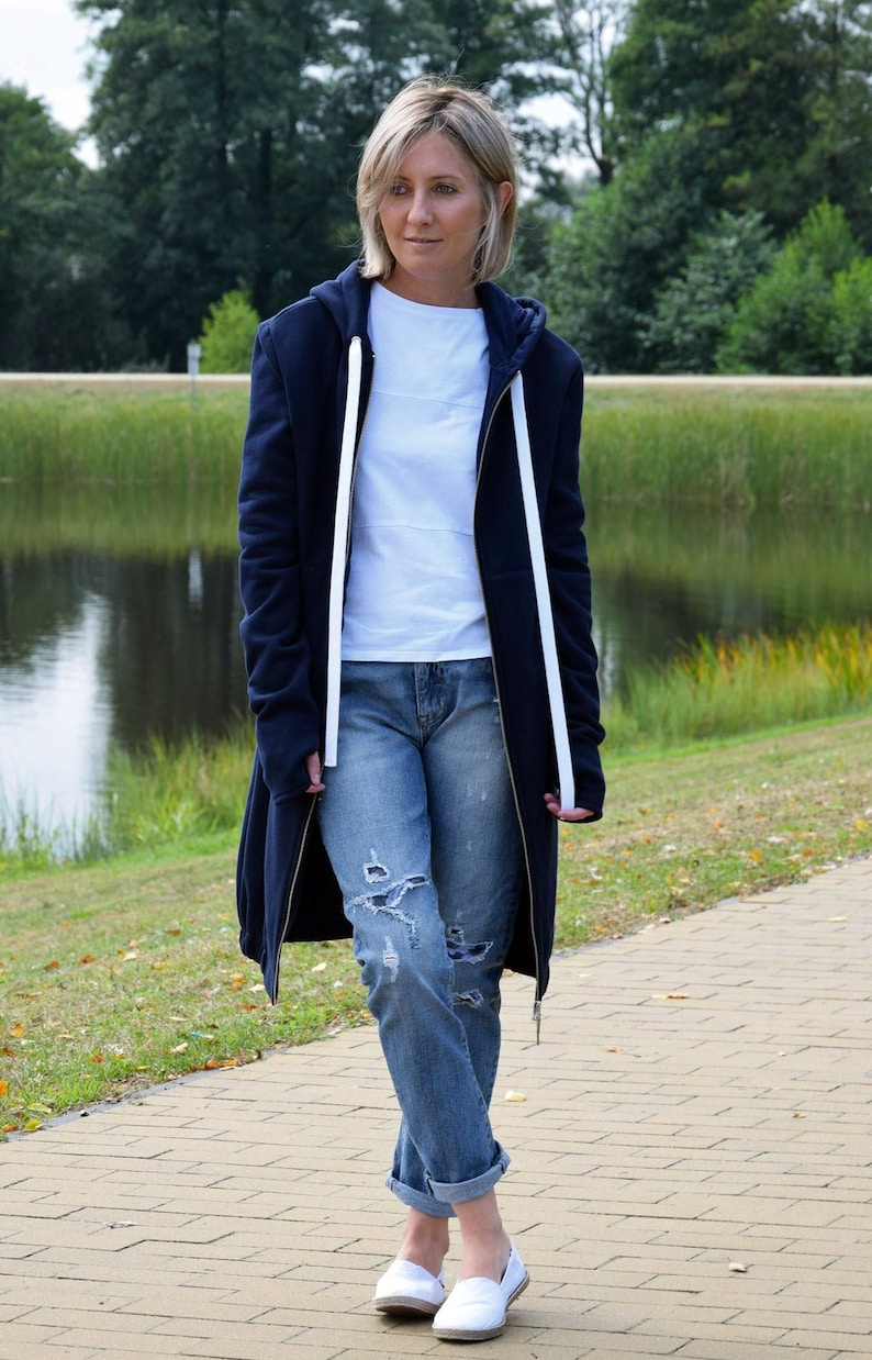May include: A woman wearing a long, dark blue hooded sweatshirt with white drawstrings, ripped light blue jeans, and white canvas sneakers.