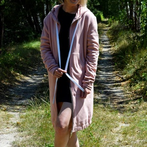 May include: A pink hooded sweatshirt with a long zipper and white drawstrings. The sweatshirt is worn by a person standing on a path in a forest.