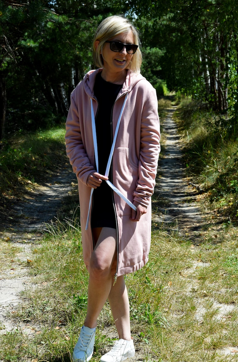 May include: A pink hooded sweatshirt with a long zipper and white drawstrings. The sweatshirt is worn by a person standing on a path in a forest.
