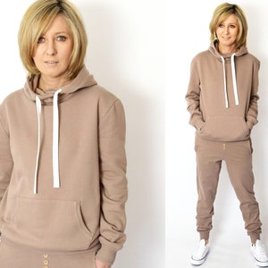 Pu&ograve; includere: Una felpa con cappuccio marrone e pantaloni della tuta abbinati con un cordoncino bianco e bottoni dorati. La modella indossa l'outfit e scarpe da ginnastica bianche.