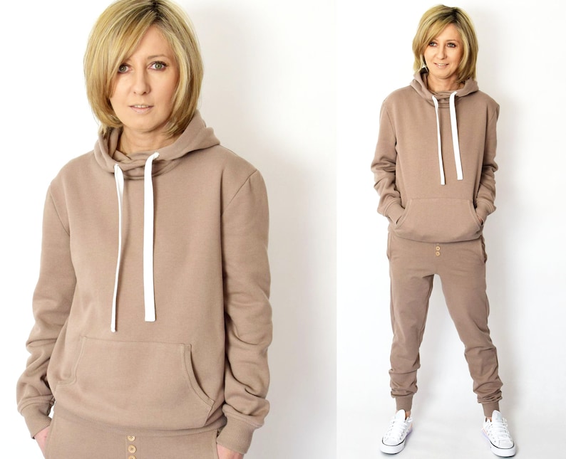 Pu&ograve; includere: Una felpa con cappuccio marrone e pantaloni della tuta abbinati con un cordoncino bianco e bottoni dorati. La modella indossa l'outfit e scarpe da ginnastica bianche.