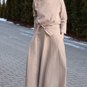 May include: A beige dress with long sleeves and a matching tie belt. The dress has a round neckline and a full skirt. The person is wearing black boots and sunglasses. The background features green trees and a paved walkway.